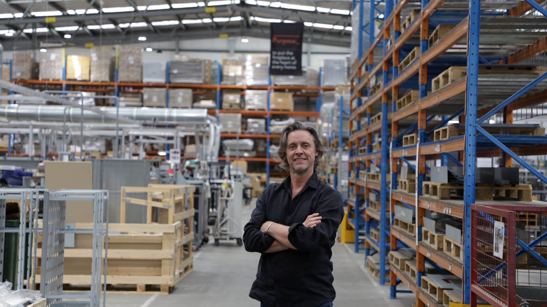 Man in black shirt stands smiling with arms crossed in a fireplace factory, surrounded by shelves and industrial equipment. A sign above reads: "Purpose: To put the fireplace at the heart of every home."
