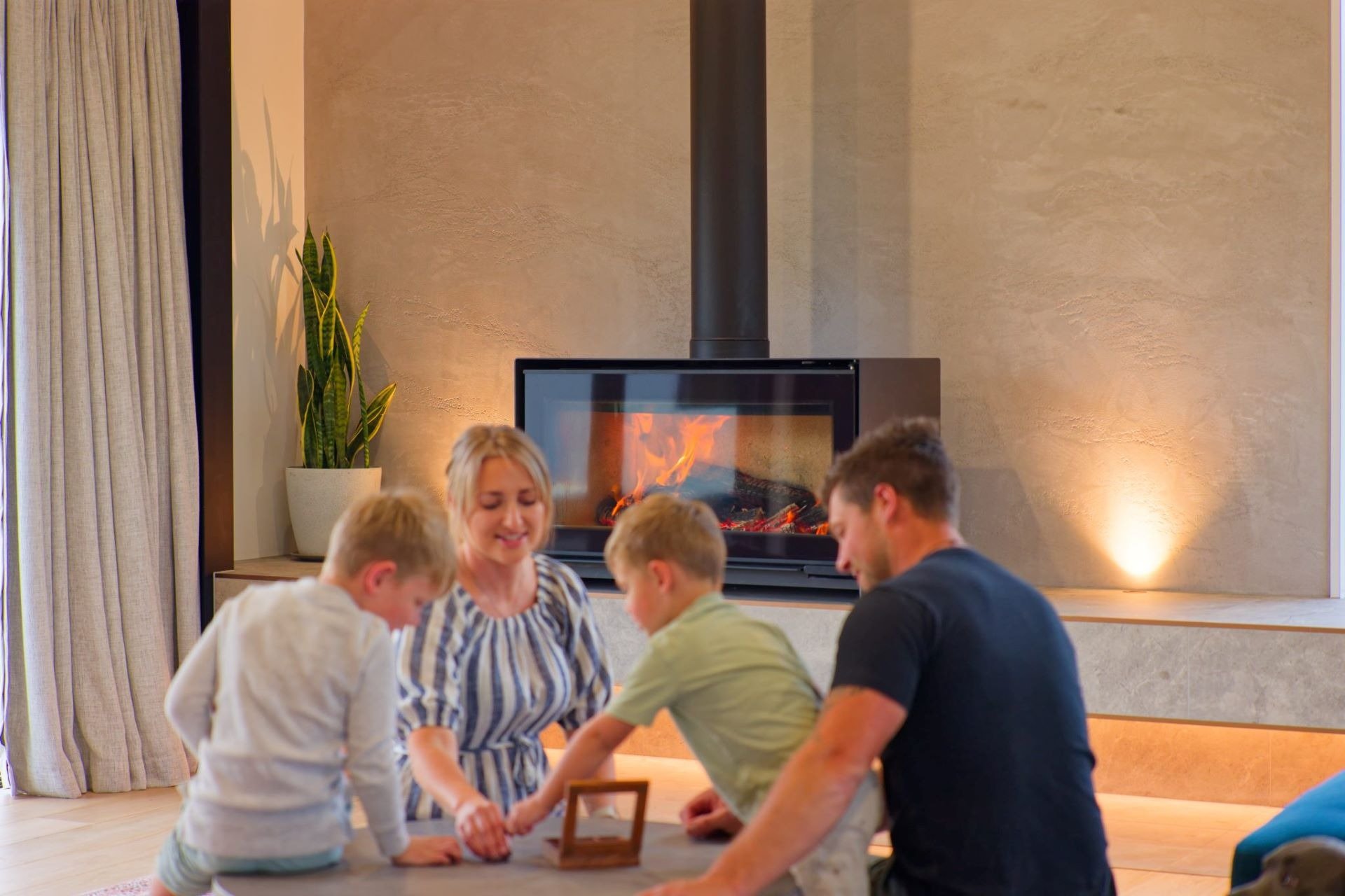 The Steele family playing together near the Escea fireplace, with a potted plant and built-in hearth bench completing the warm, modern interior.